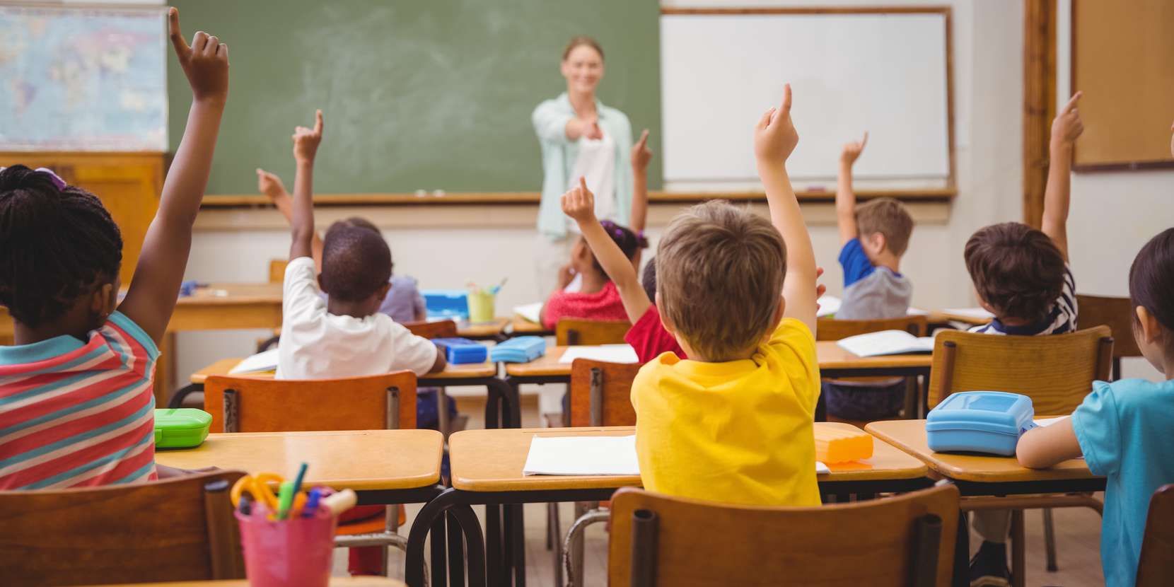 classroom full of children raising their hands up with a teacher in the background